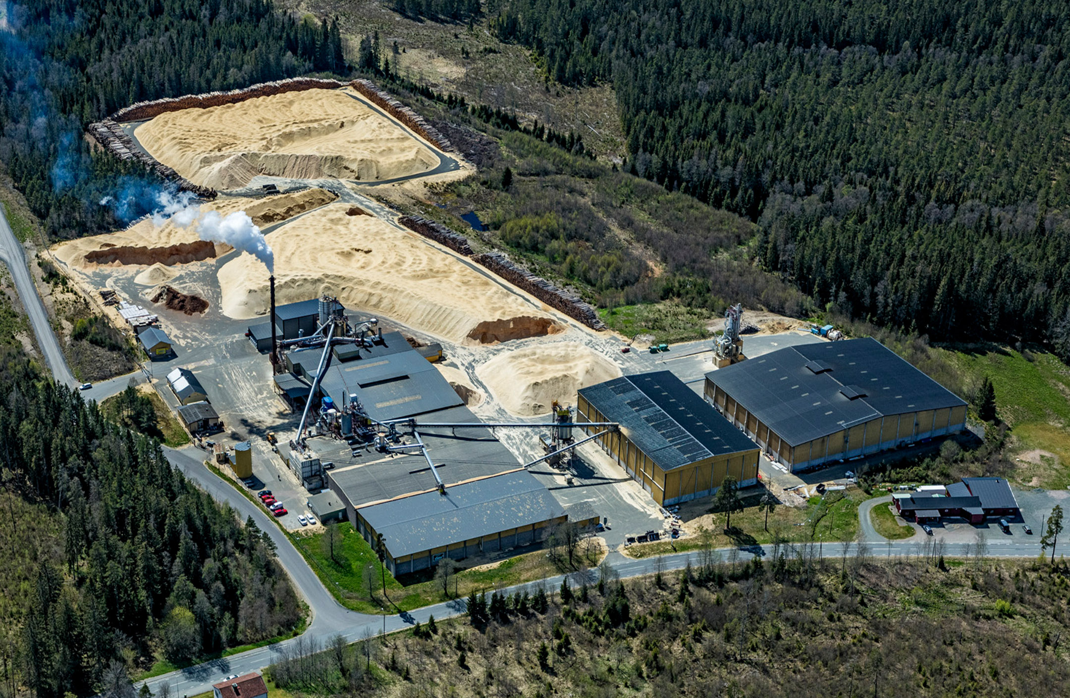 Flygbild pelletsfabriken i Malmbäck. Byggnader, spånhögar, skog och vattenånga från torkprocessen