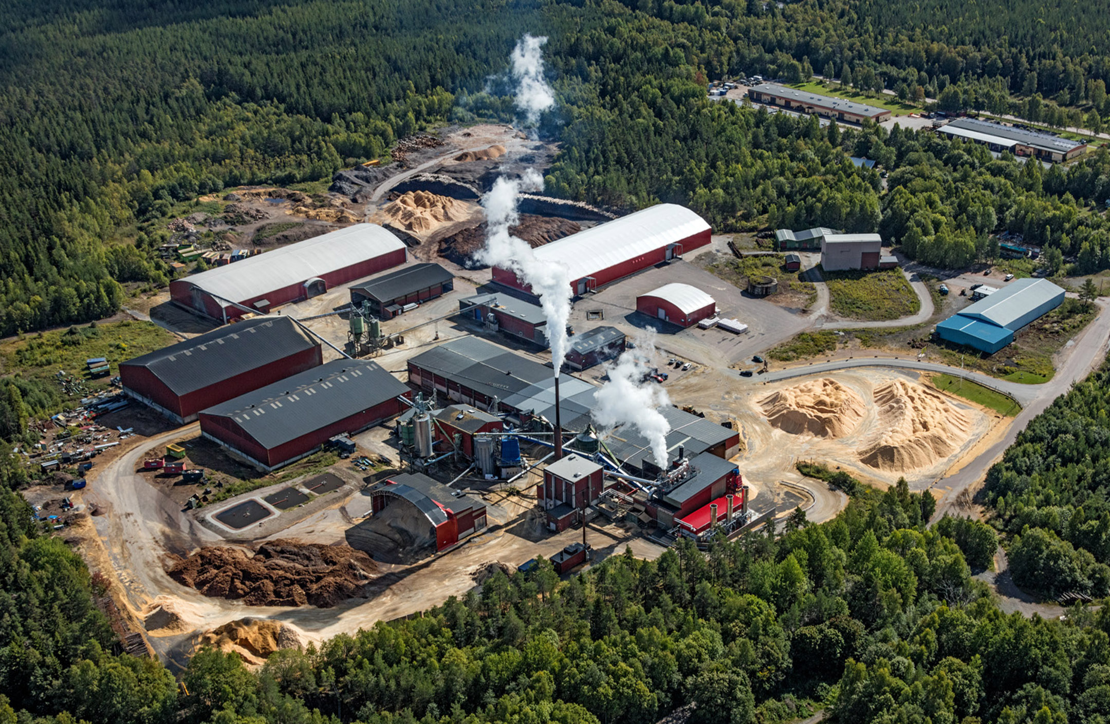 Flygbild pelletsfabriken i Norberg. Byggnader, spånhögar, skog och vattenånga från torkprocessen