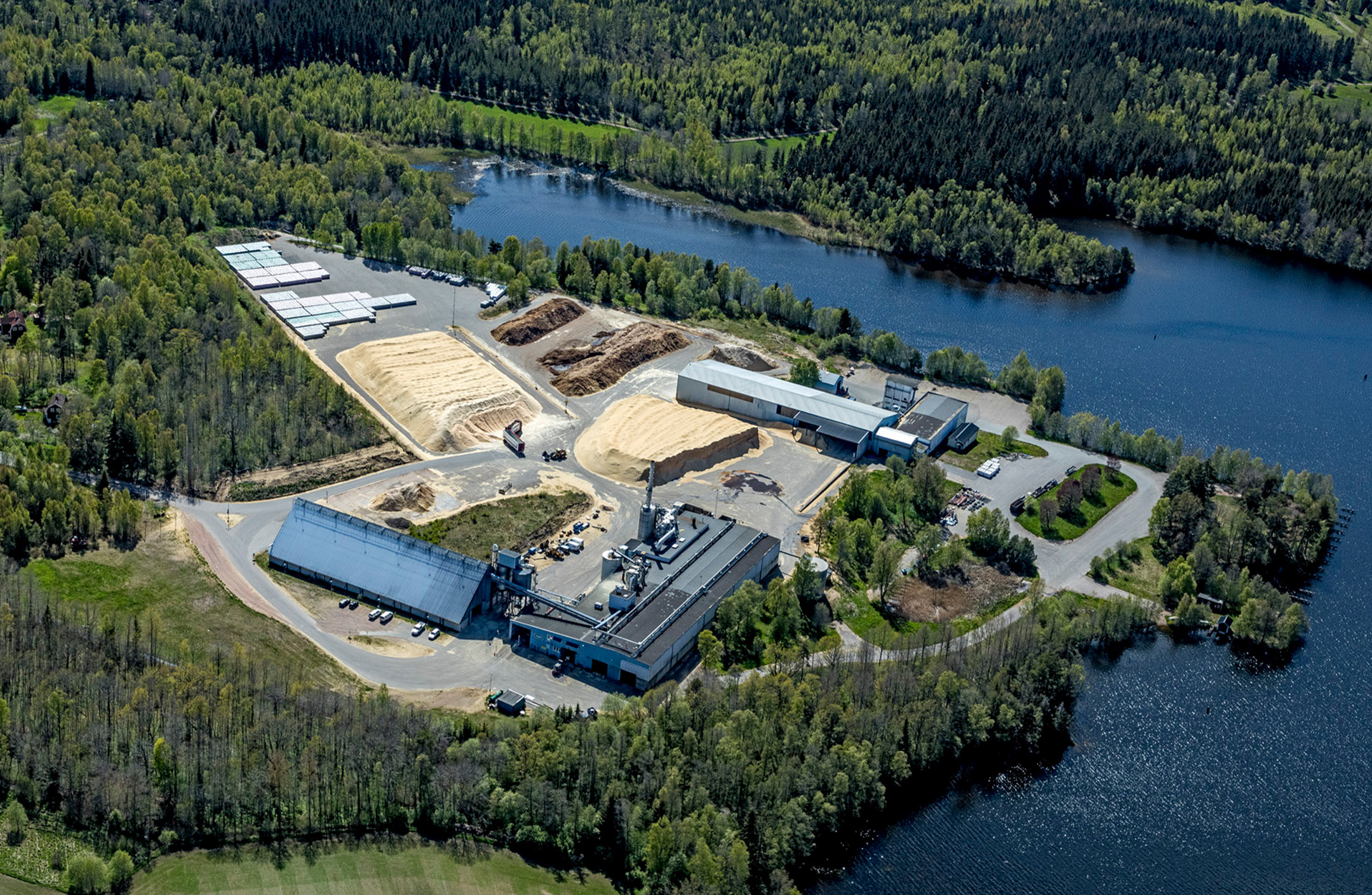 Flygbild pelletsfabriken i Norberg. Byggnader, spånhögar på asfaltplan, omgivet av vatten och skog