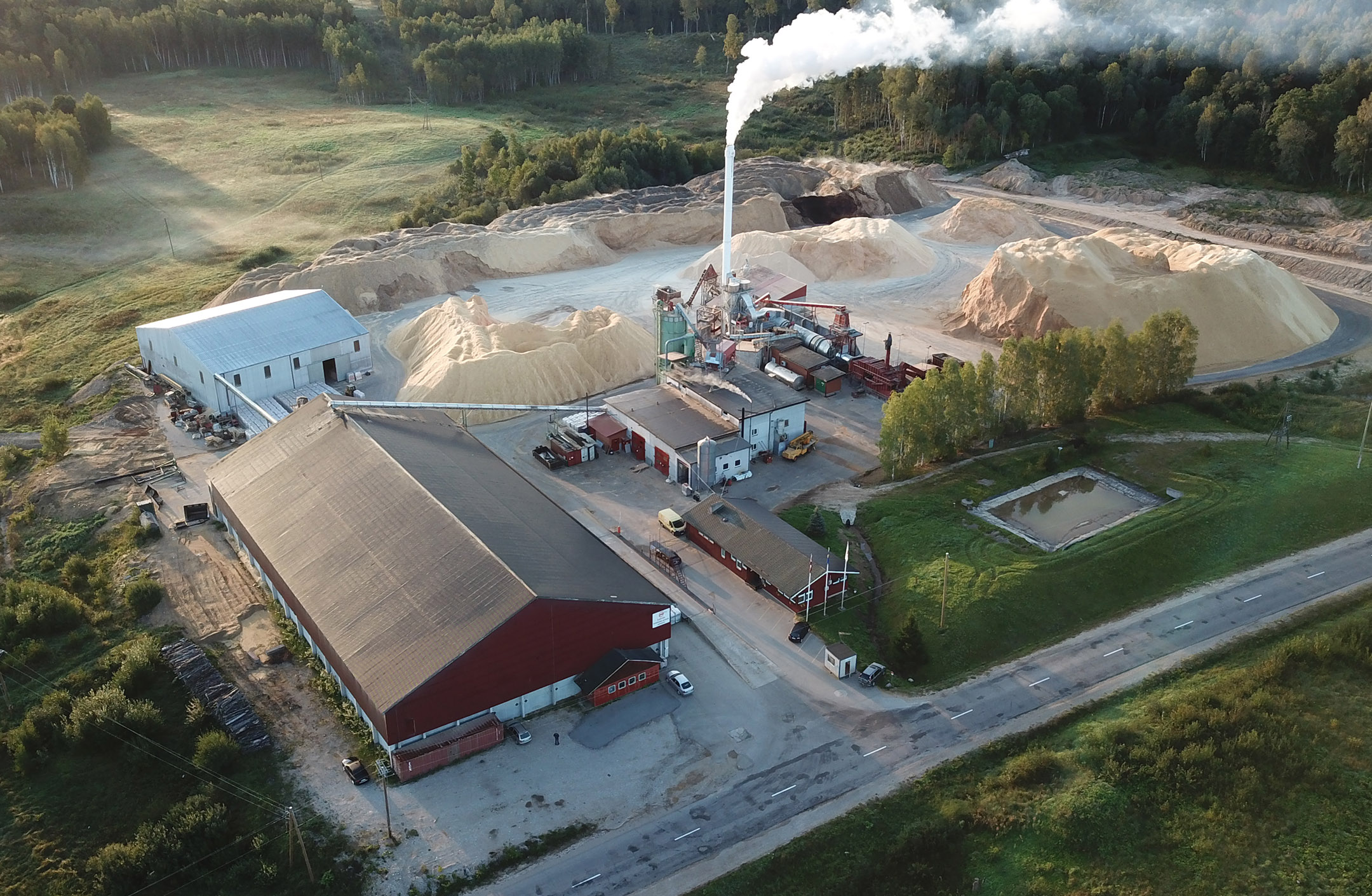 Flygbild pelletsfabriken i Talsi, Lettland. Byggnader, spånhögar och vattenånga från torkprocessen