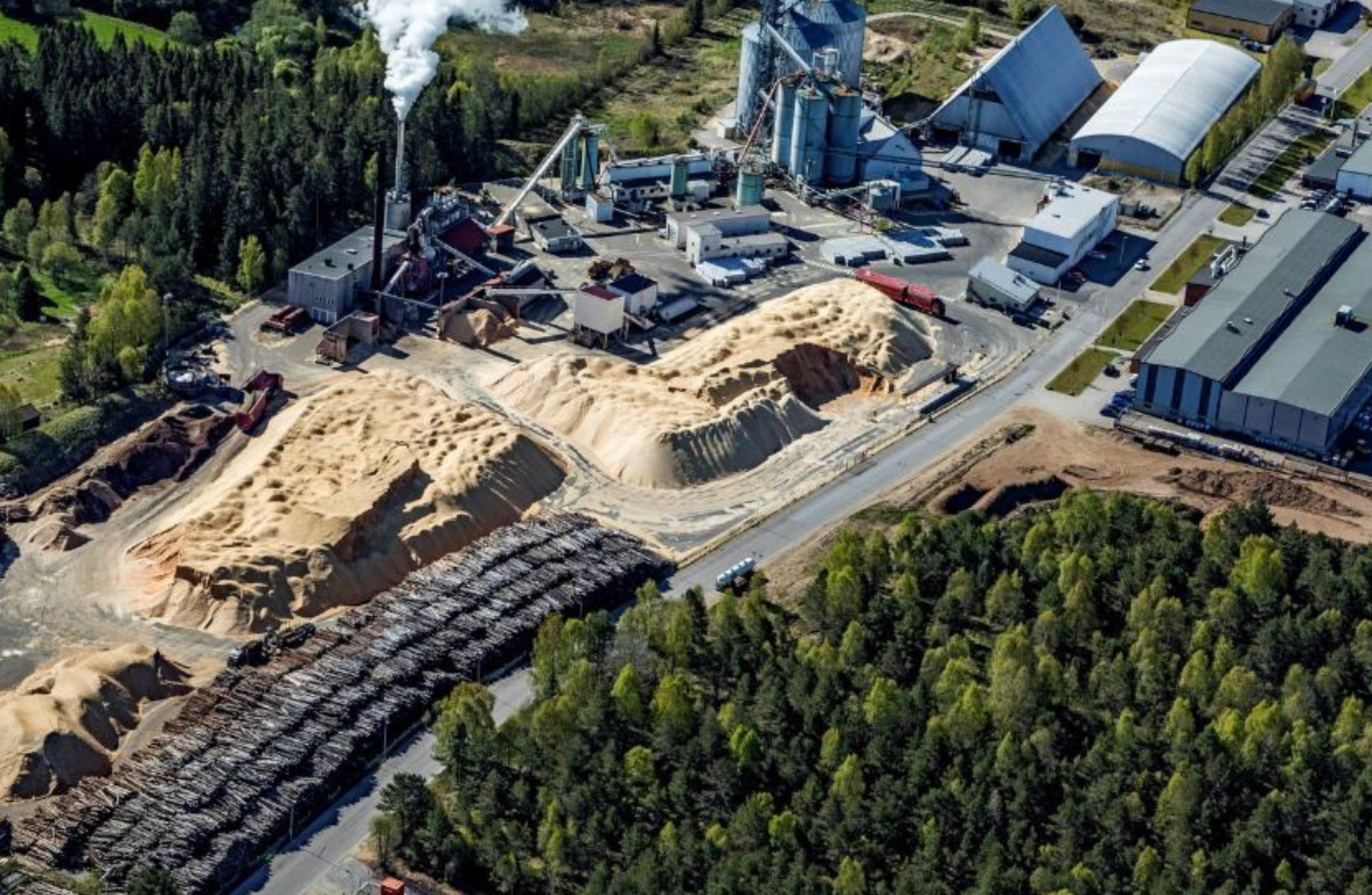 Flygbild pelletsfabriken och fjärrvärmepanna i Ulricehamn. Byggnader, spånhögar, skog och vattenånga från torkprocessen